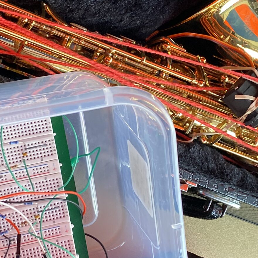 Close-up of the EMG saxophone build, with the breadboarded acquisition circuit and its jumper wires in the foreground and the wired saxophone in its case behind it